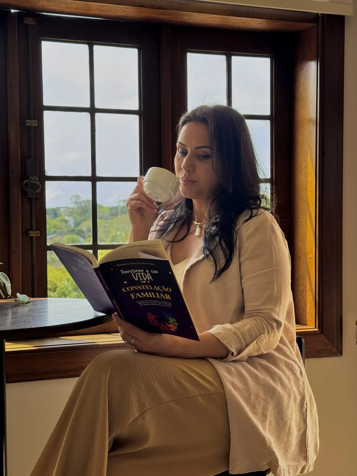 Roberta Dias lendo à janela, com uma xícara na mão
