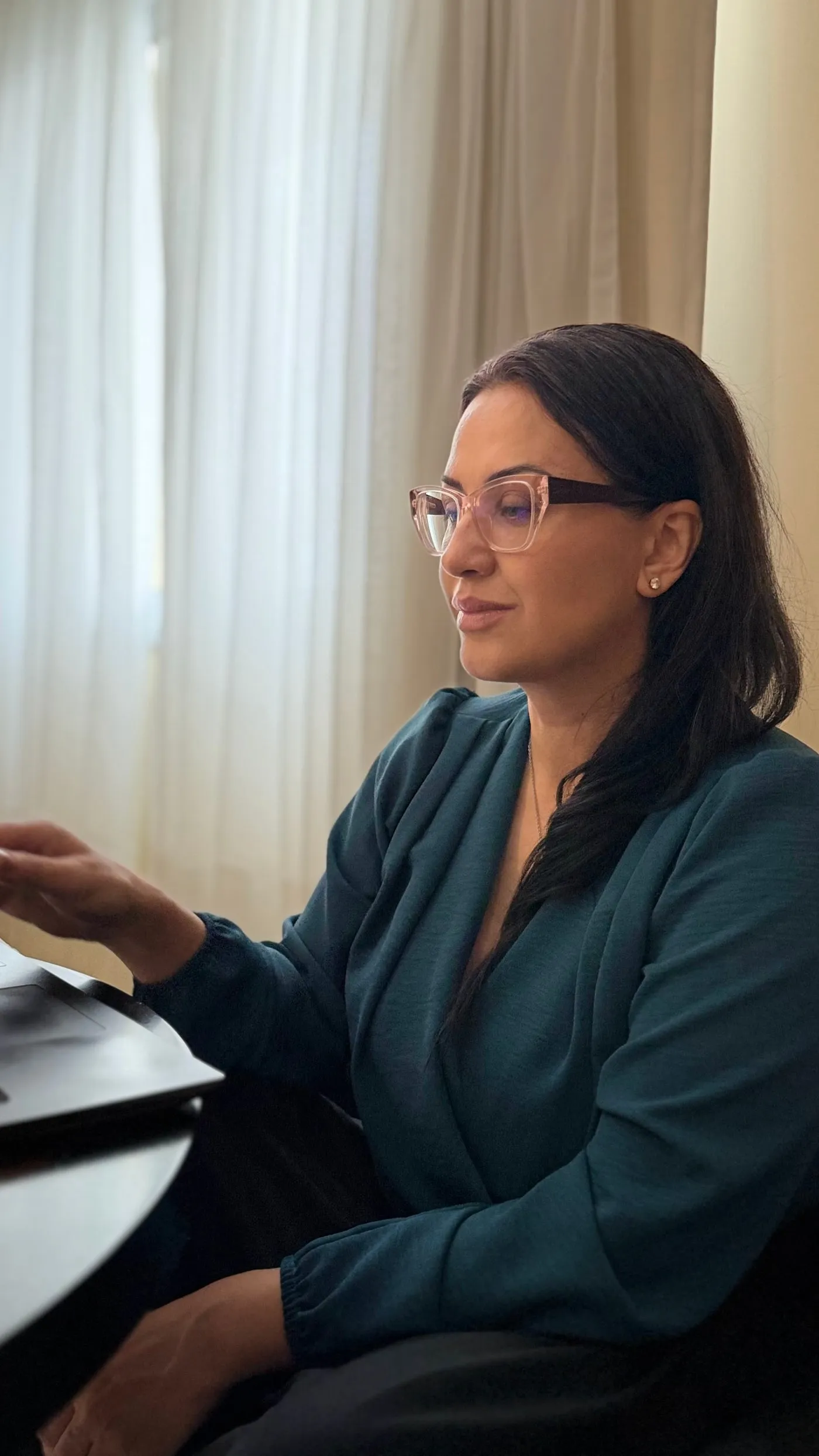 Roberta Dias de óculos, trabalhando no notebook perto da janela